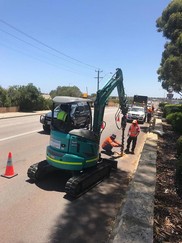 A man is kneeling next to a green excavator on the side of the road.