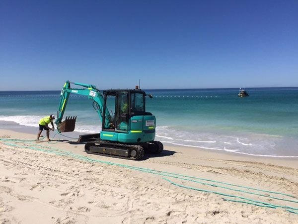 A blue excavator is parked on the beach next to the ocean.