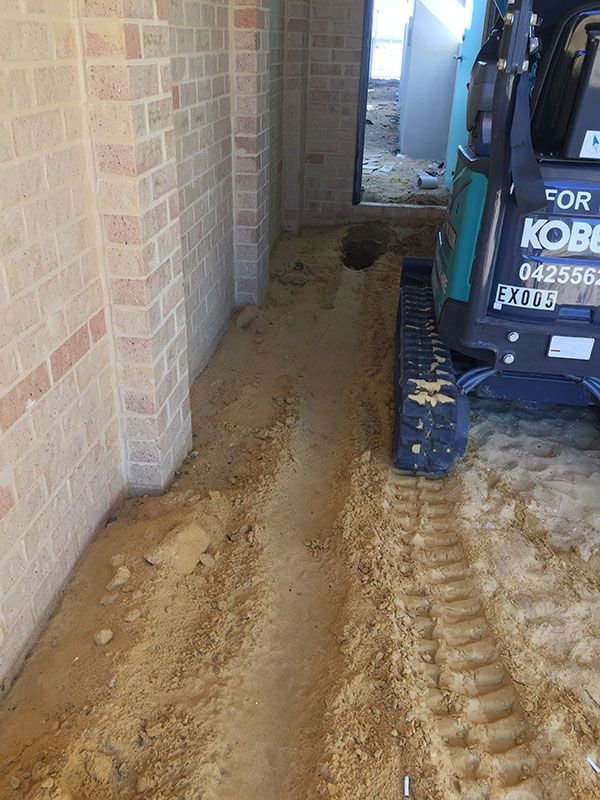 A small excavator is digging a hole in the ground next to a brick wall.