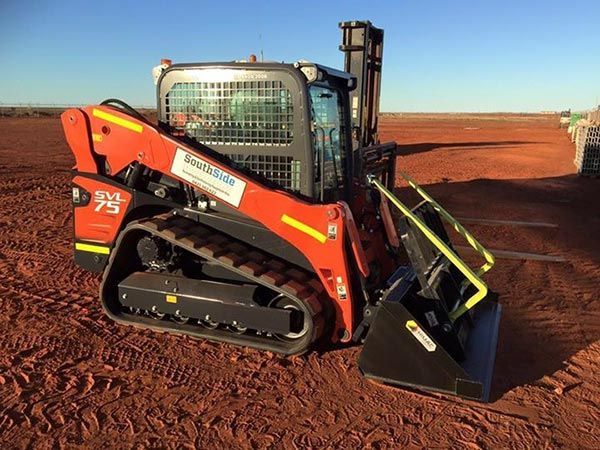 A skid steer with a forklift attached to it is parked in a dirt field.