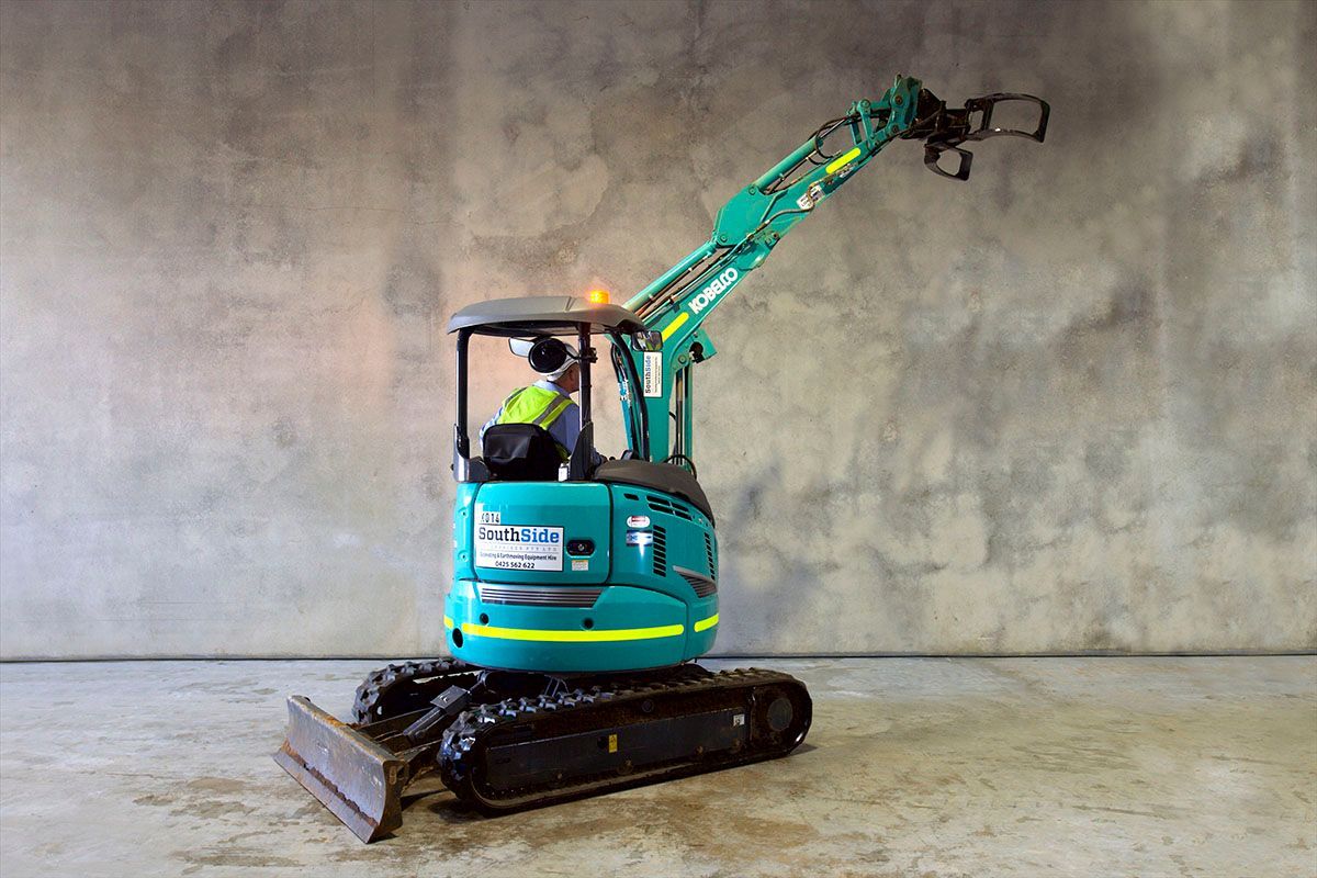 A small blue excavator is parked in front of a concrete wall.