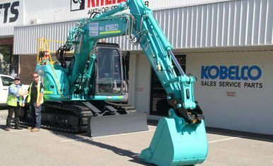 A blue excavator is parked in front of a kobelco sales service parts store.