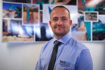 A man in a blue shirt and tie is standing in front of a wall with pictures on it.