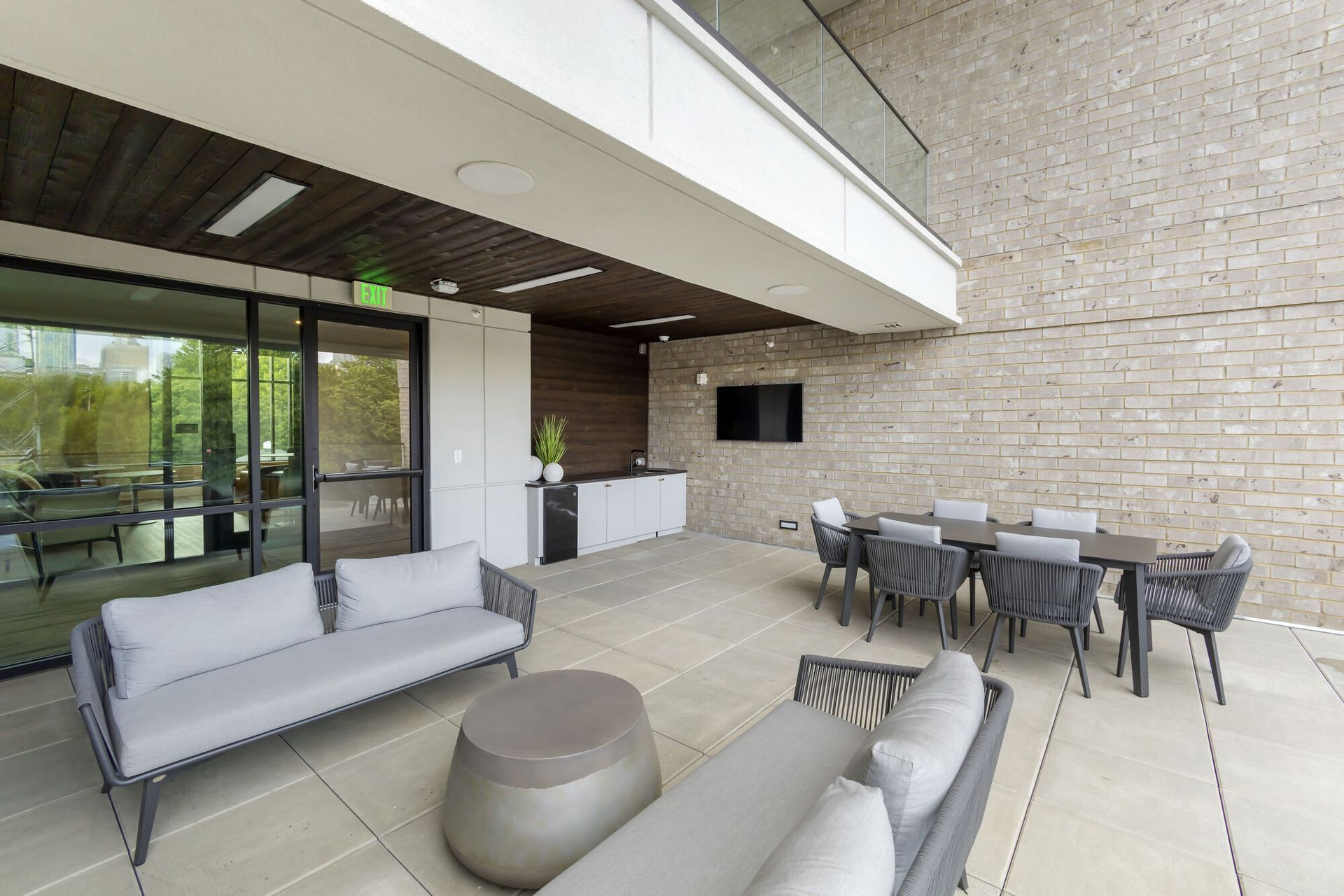 Outdoor community lounge with gray sectional seating, dining table, kitchenette, and wall-mounted TV under a covered patio.