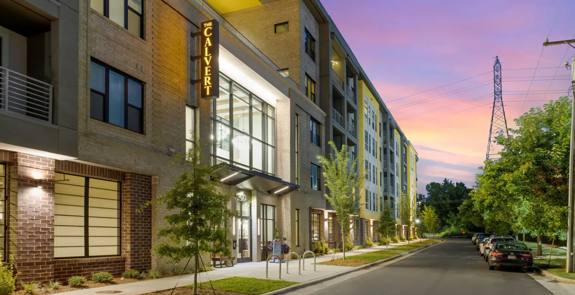 Exterior view of a modern apartment building with an illuminated entrance and signage at dusk.