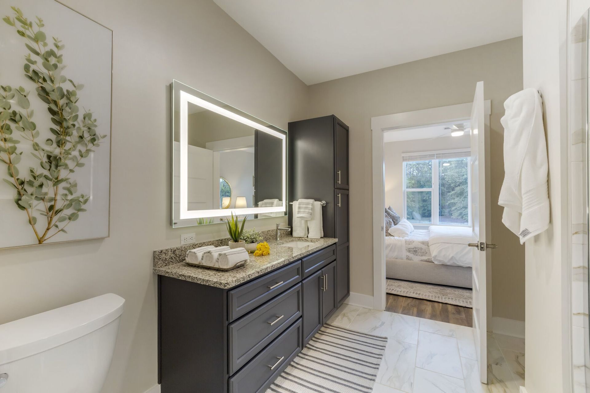Modern bathroom with dark cabinetry, granite counter, backlit mirror, and doorway to a bedroom.