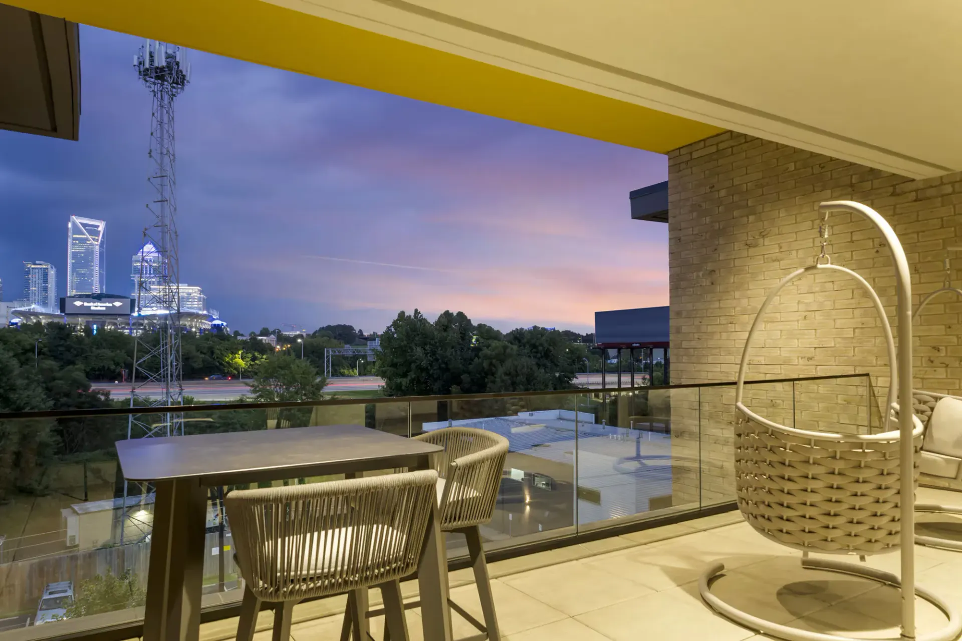 Outdoor balcony with a square table, two woven chairs, and a hanging chair; city skyline at dusk.
