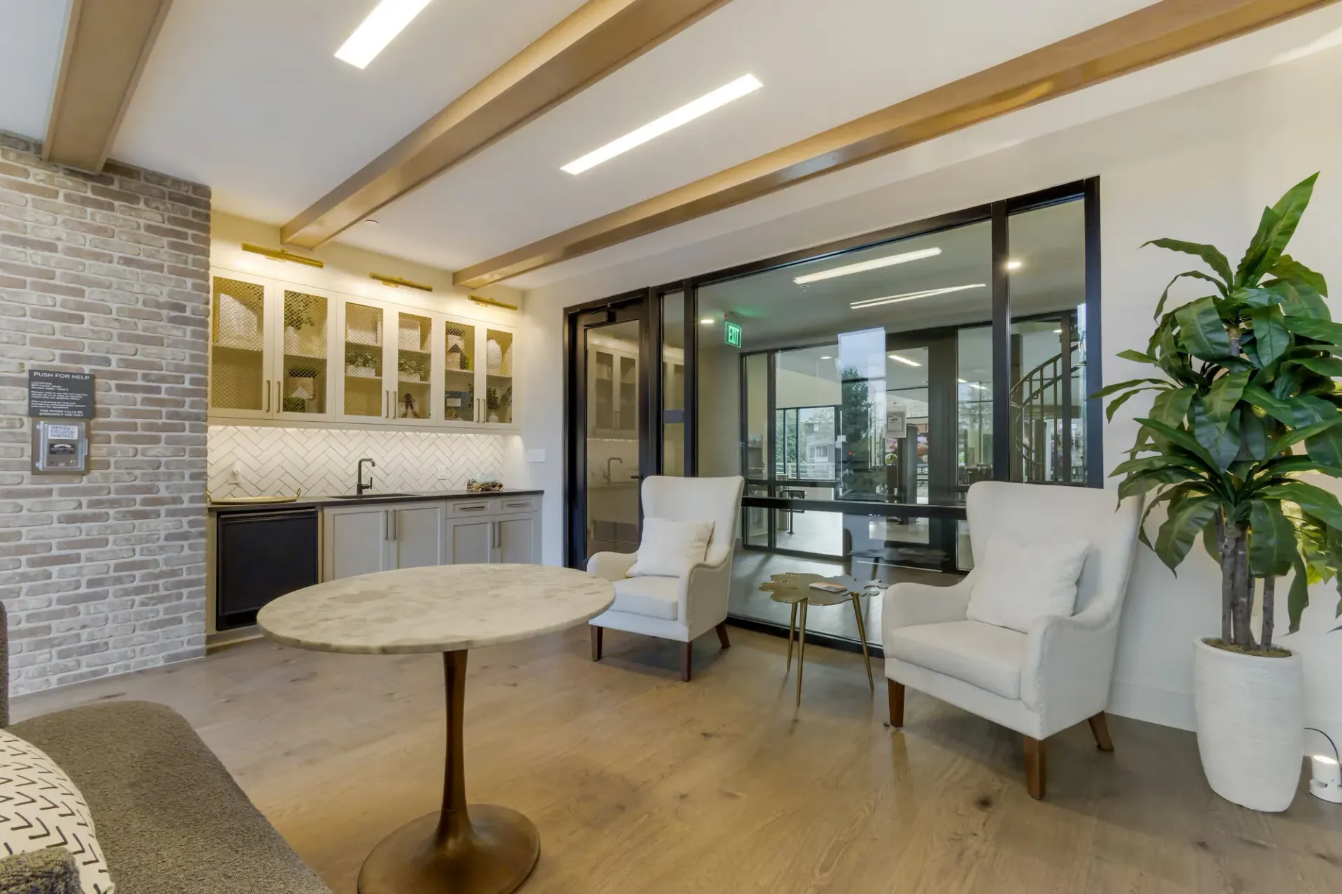 Modern community lounge with kitchenette, brick wall, and glass doors to the outdoors.