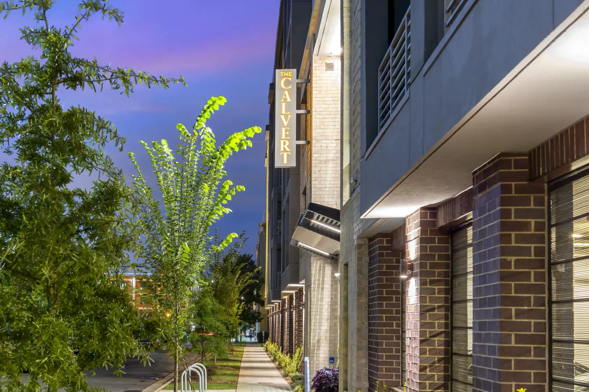 Exterior view of a modern apartment building at dusk with a lit sign and trees along the sidewalk