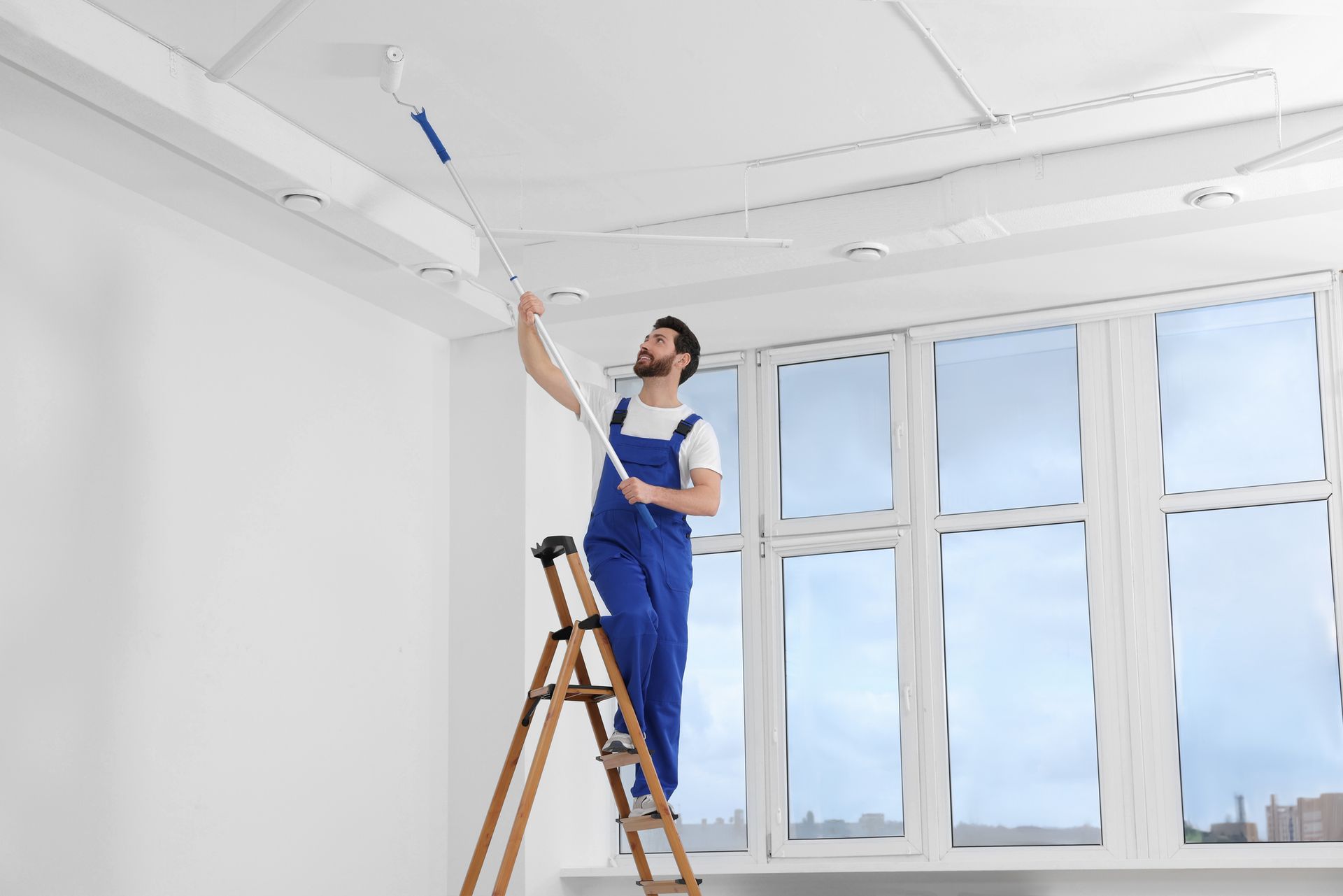 A man is standing on a ladder painting the ceiling of a room.