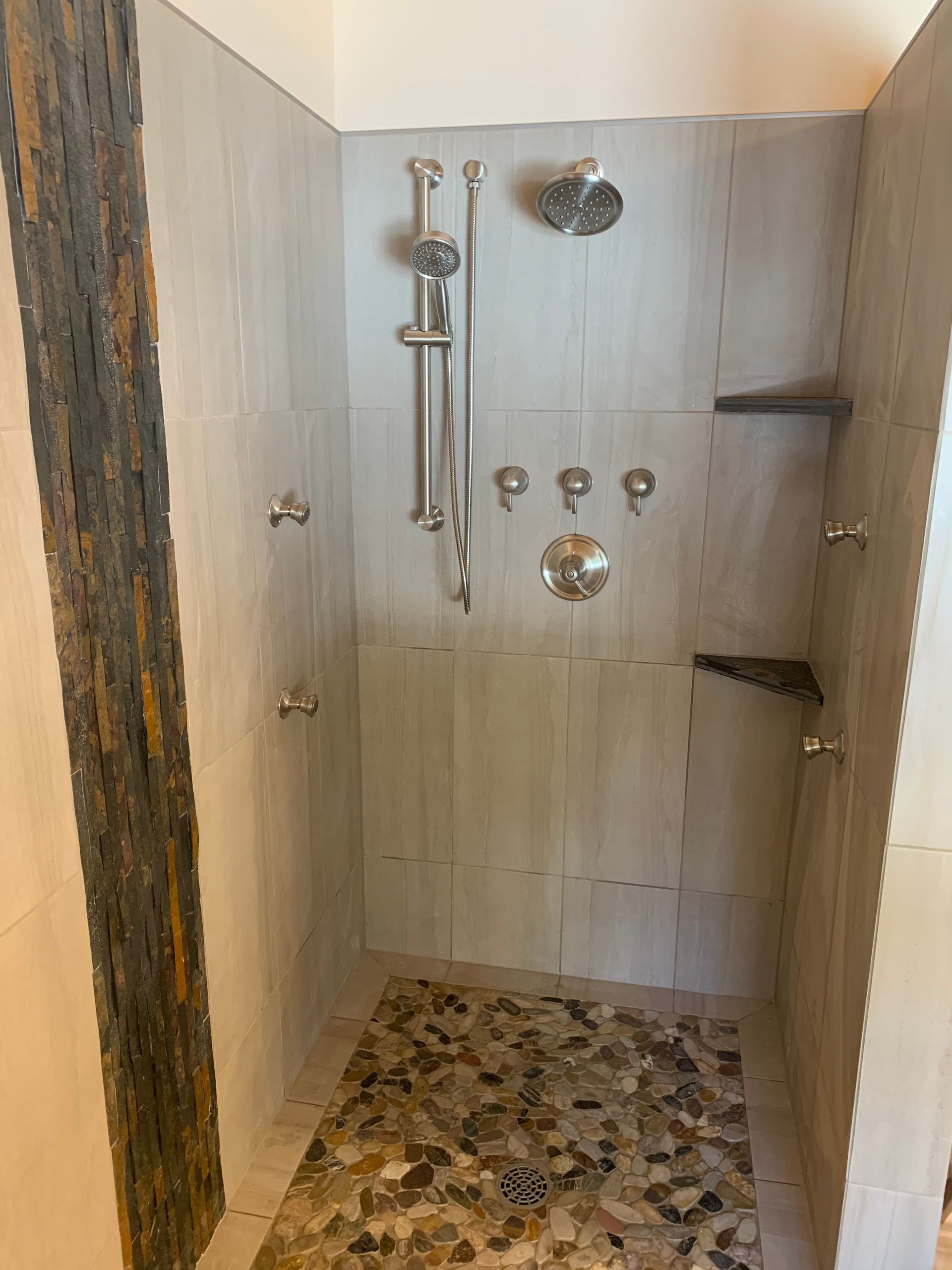 Shower with pebble tile floor, light gray walls, and multiple silver showerheads and fixtures.