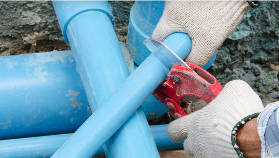 Hands wearing gloves cutting blue PVC pipes with a saw in an outdoor setting.