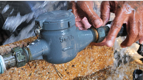 Hands tightening a fitting on a blue water meter, water gushing out.