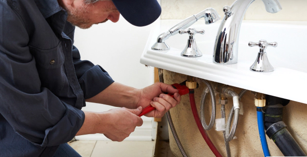 Plumber in blue shirt and cap uses pliers to work on pipes under a white bathtub.