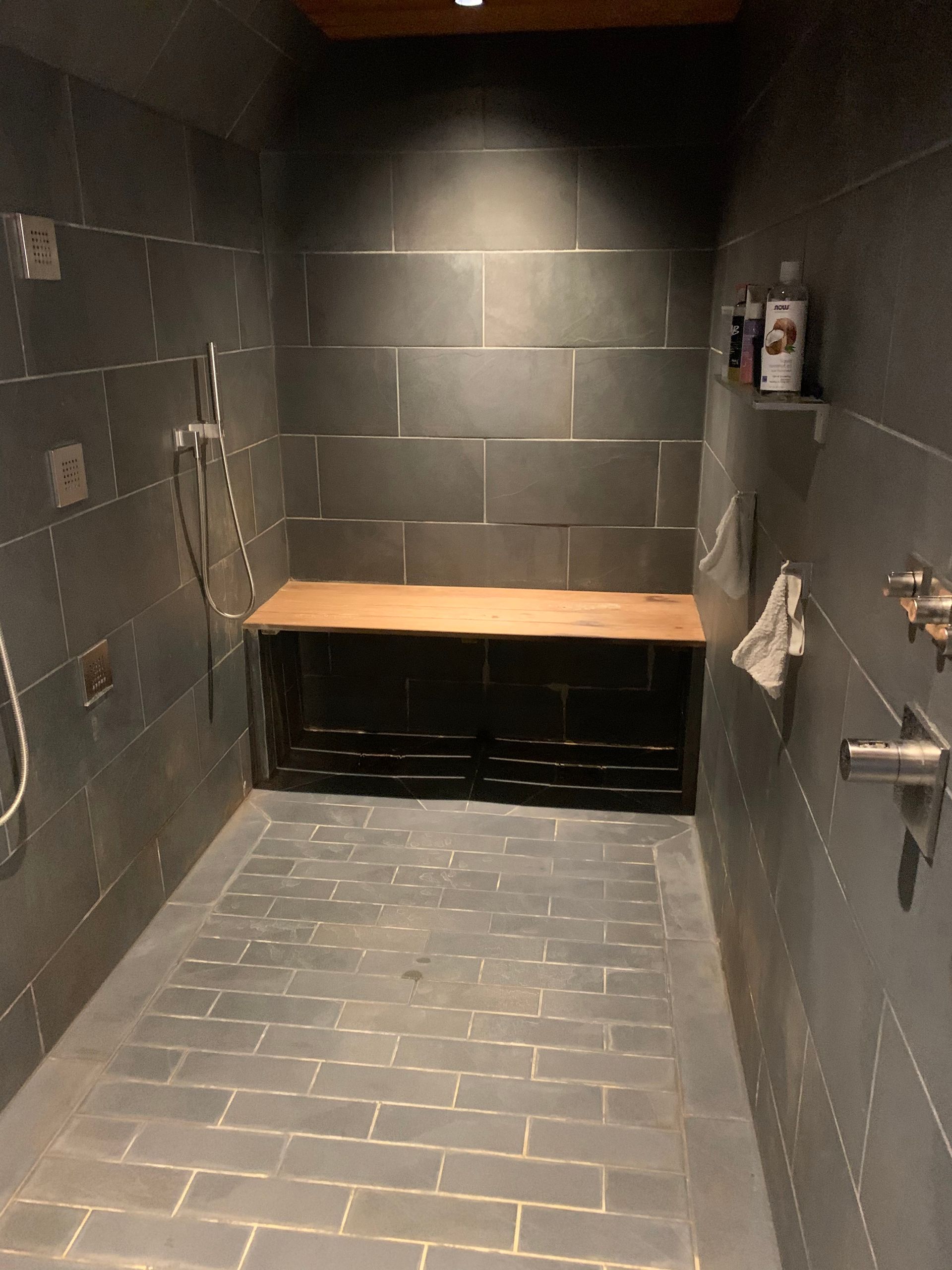 Dark tile shower with wooden bench, toiletries on a shelf, and shower controls.