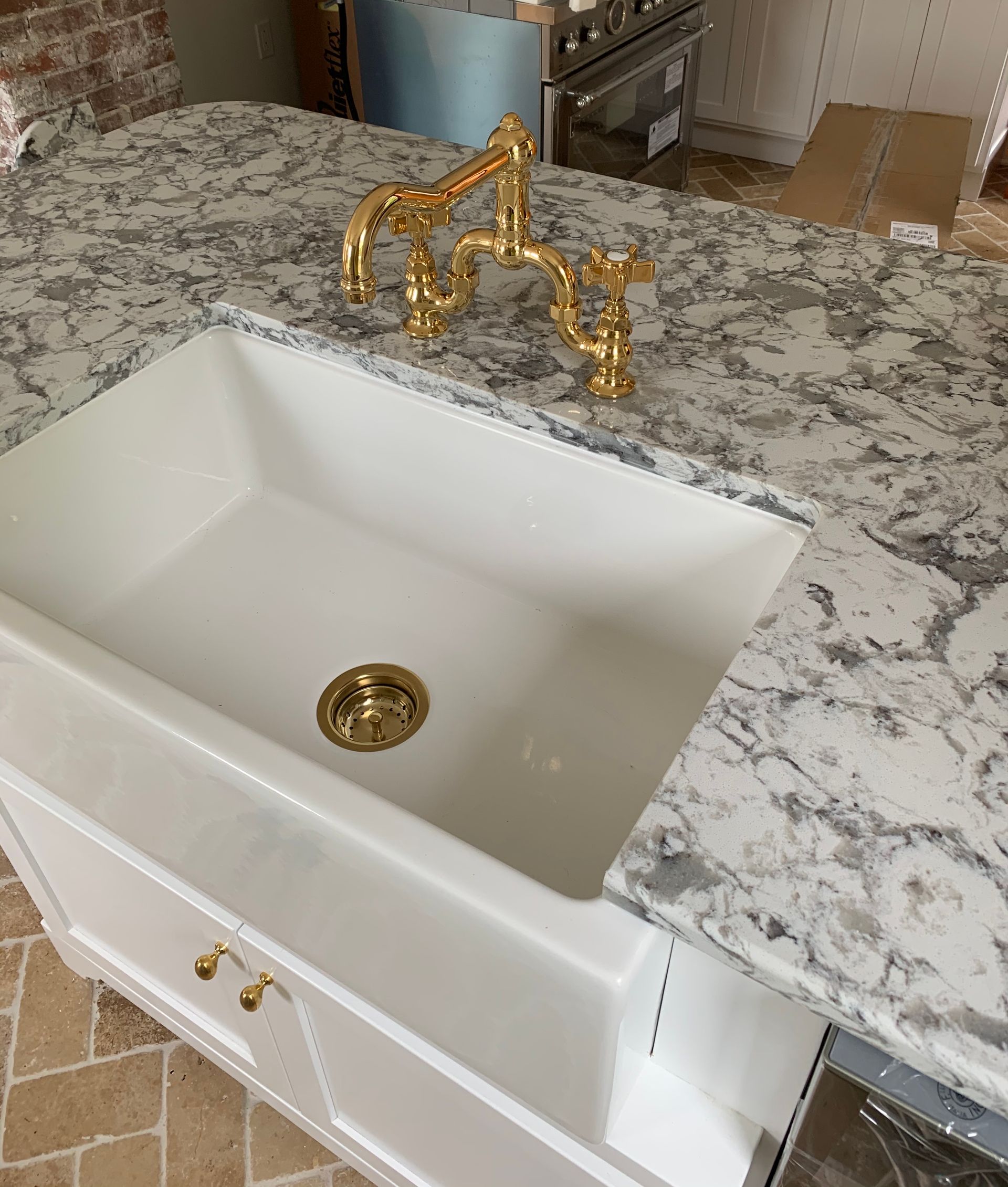 White farmhouse sink with gold faucet on quartz countertop, in a kitchen.