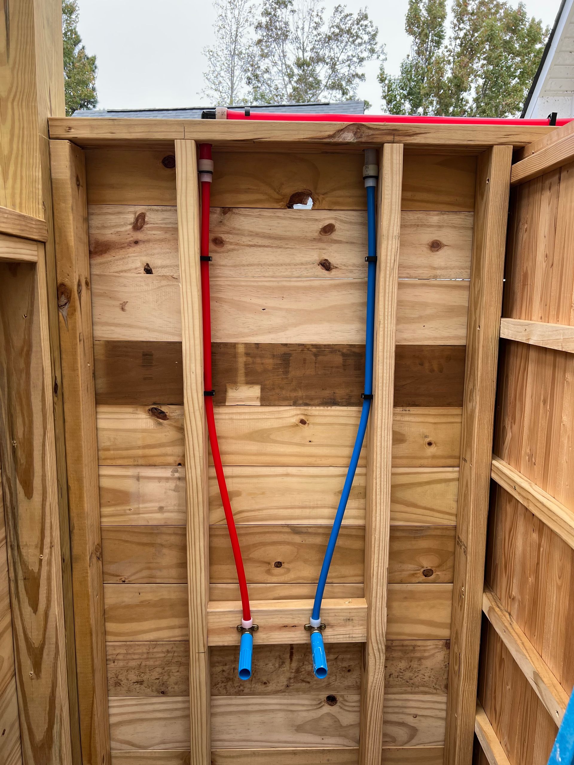 Interior view of a wooden shower stall with red and blue water pipes running vertically.