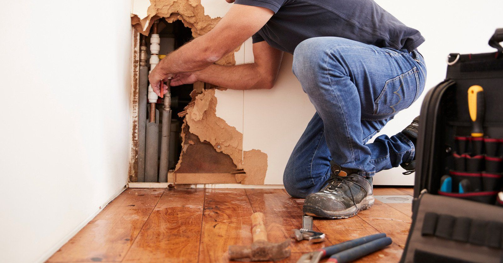 Plumber kneels, repairing pipes inside a wall; a toolbox sits nearby.
