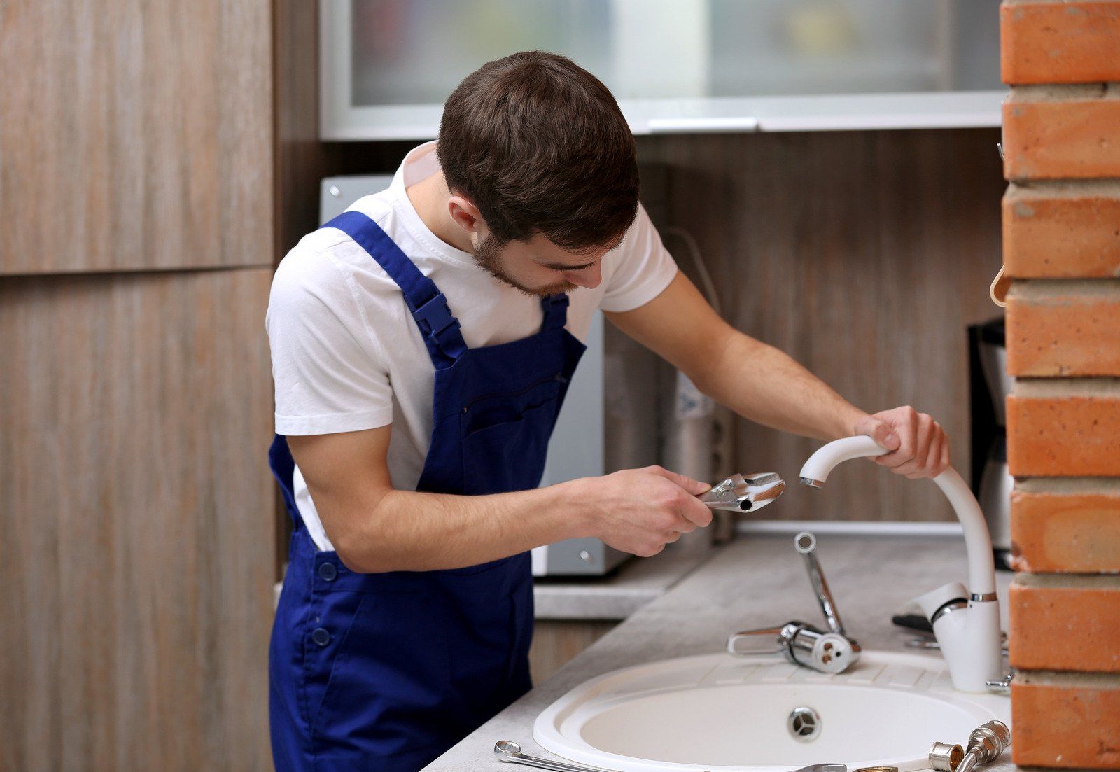 Plumber in blue overalls repairing a kitchen faucet near a sink and brick wall.