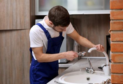 Plumber in blue overalls repairing a kitchen faucet near a sink and brick wall.