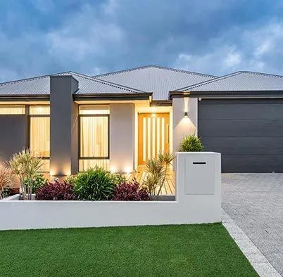 Modern single-story house with lit windows, dark garage, white fence, and trimmed lawn at dusk