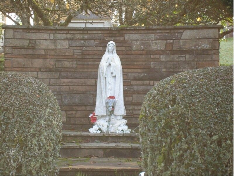 White statue of the Virgin Mary with flowers, set between trimmed bushes and stone steps in a garden shrine