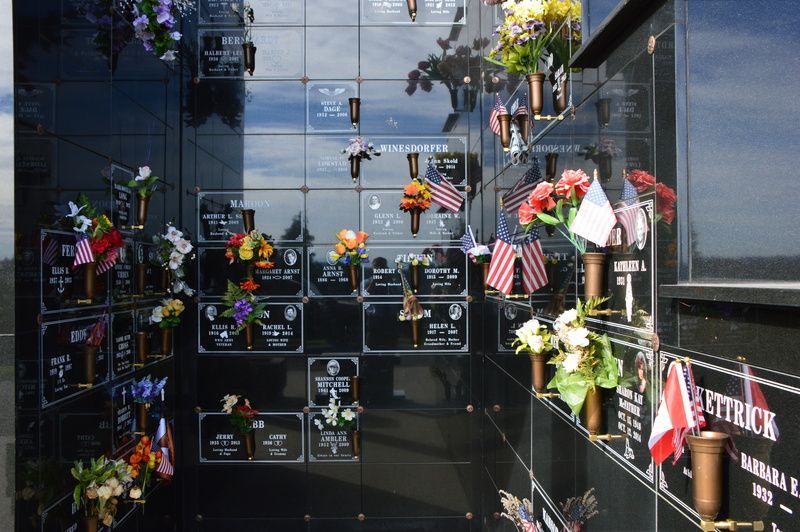 Memorial wall with many small plaques, flowers, and flags on glossy black tiles outdoors