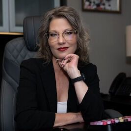 A woman wearing glasses and a black jacket is sitting at a desk with her hand on her chin.