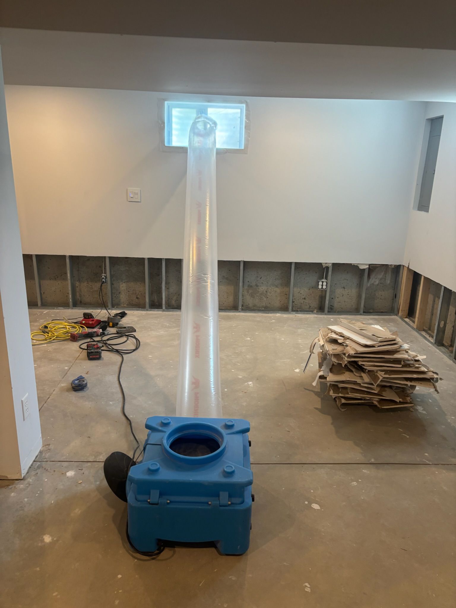 Basement being dried; a blue dehumidifier is connected to a window with clear tubing. Drywall removed from wall.