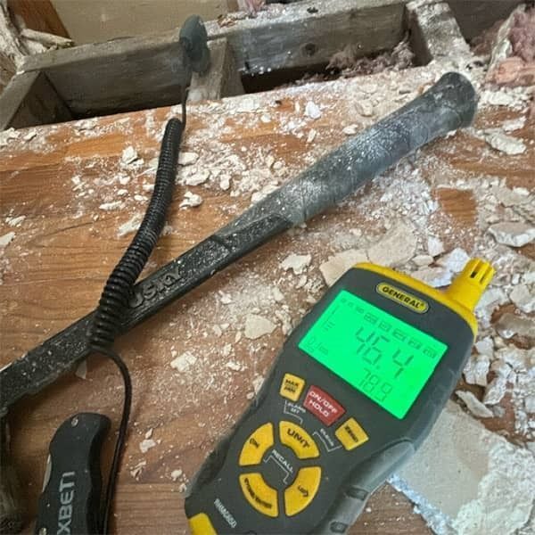 A moisture meter reads 46.4%, near a crowbar, on a dusty surface, assessing for potential water damage.