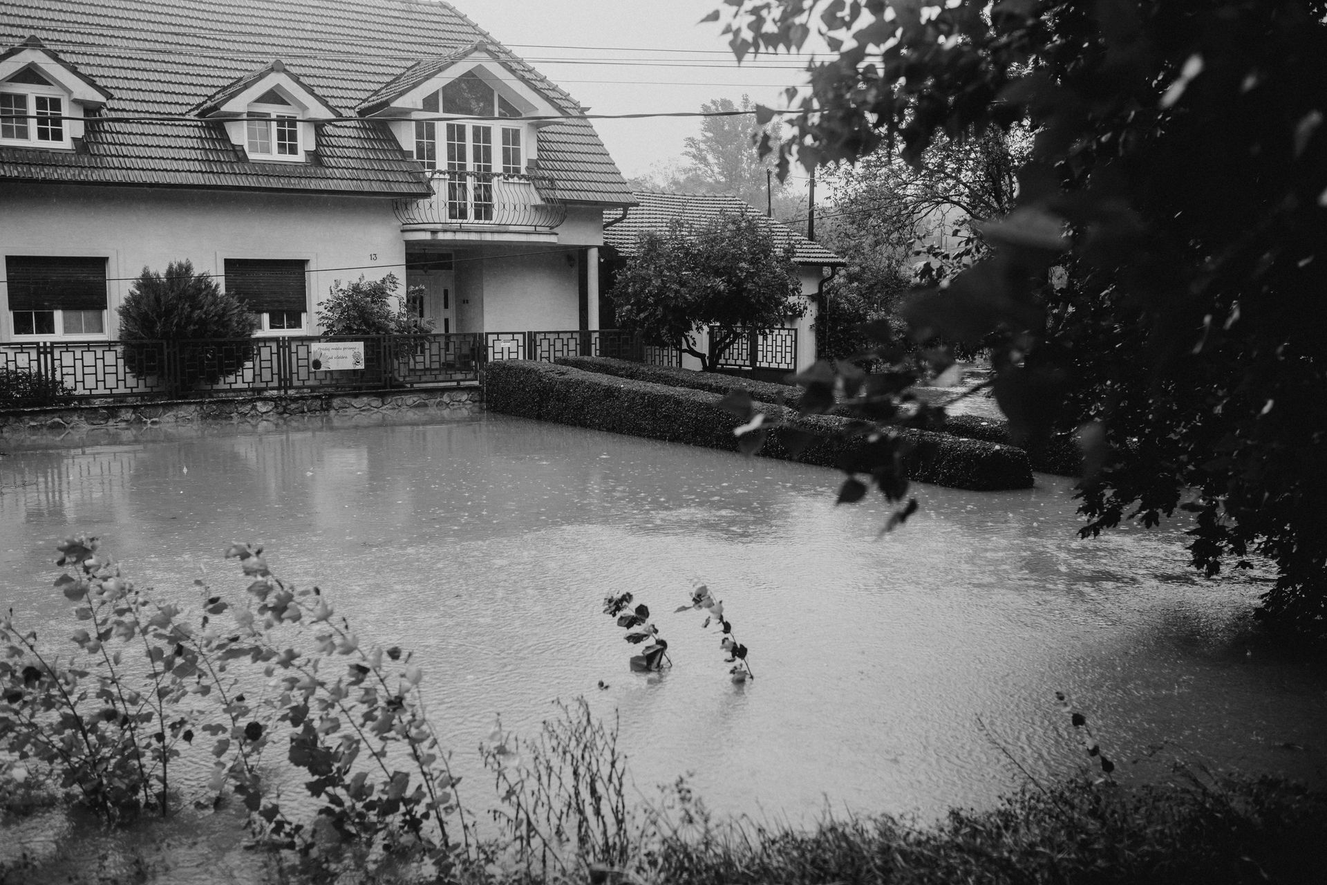 Flooded residential area; water surrounds a two-story house with dormers, bushes, and trees.