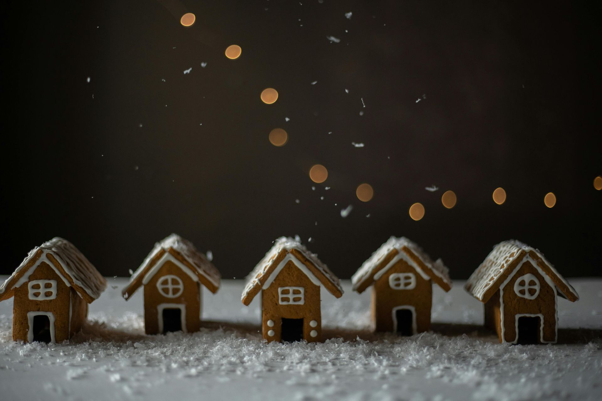 Five gingerbread houses on a snowy surface with string lights in the background.