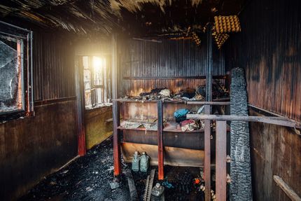 Interior of a burnt room, charred walls and furniture, debris on the floor, bright light from a window.