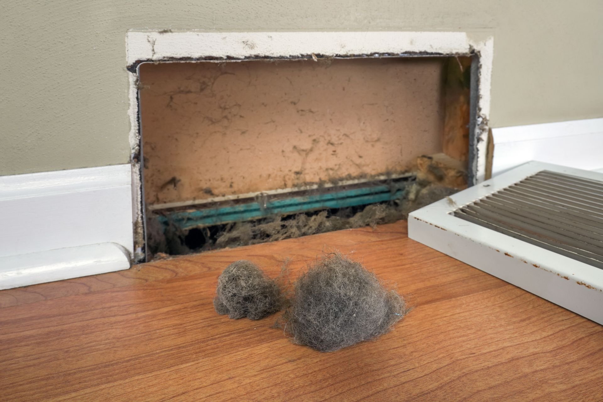A dirty air vent with debris and two dusty gray balls on a wood floor.