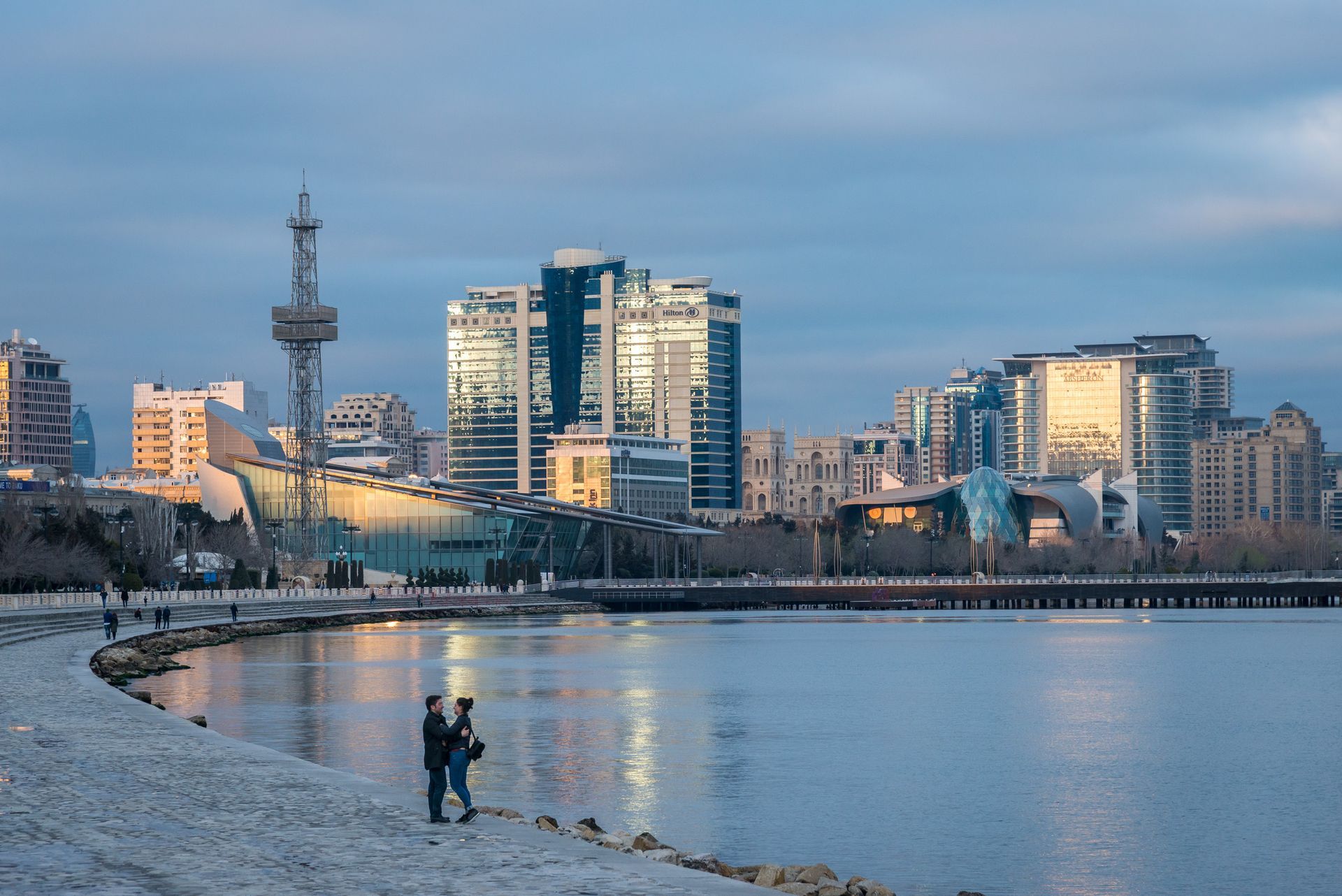 Two people are standing on the shore of a lake in front of a city skyline.