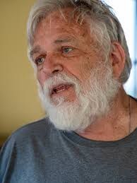 A man with a beard and gray hair is wearing a gray shirt.