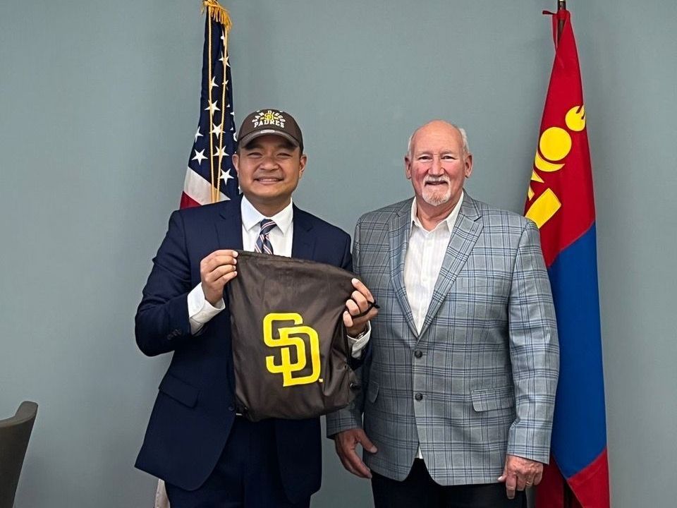Two men are standing next to each other in front of a flag holding a bag.