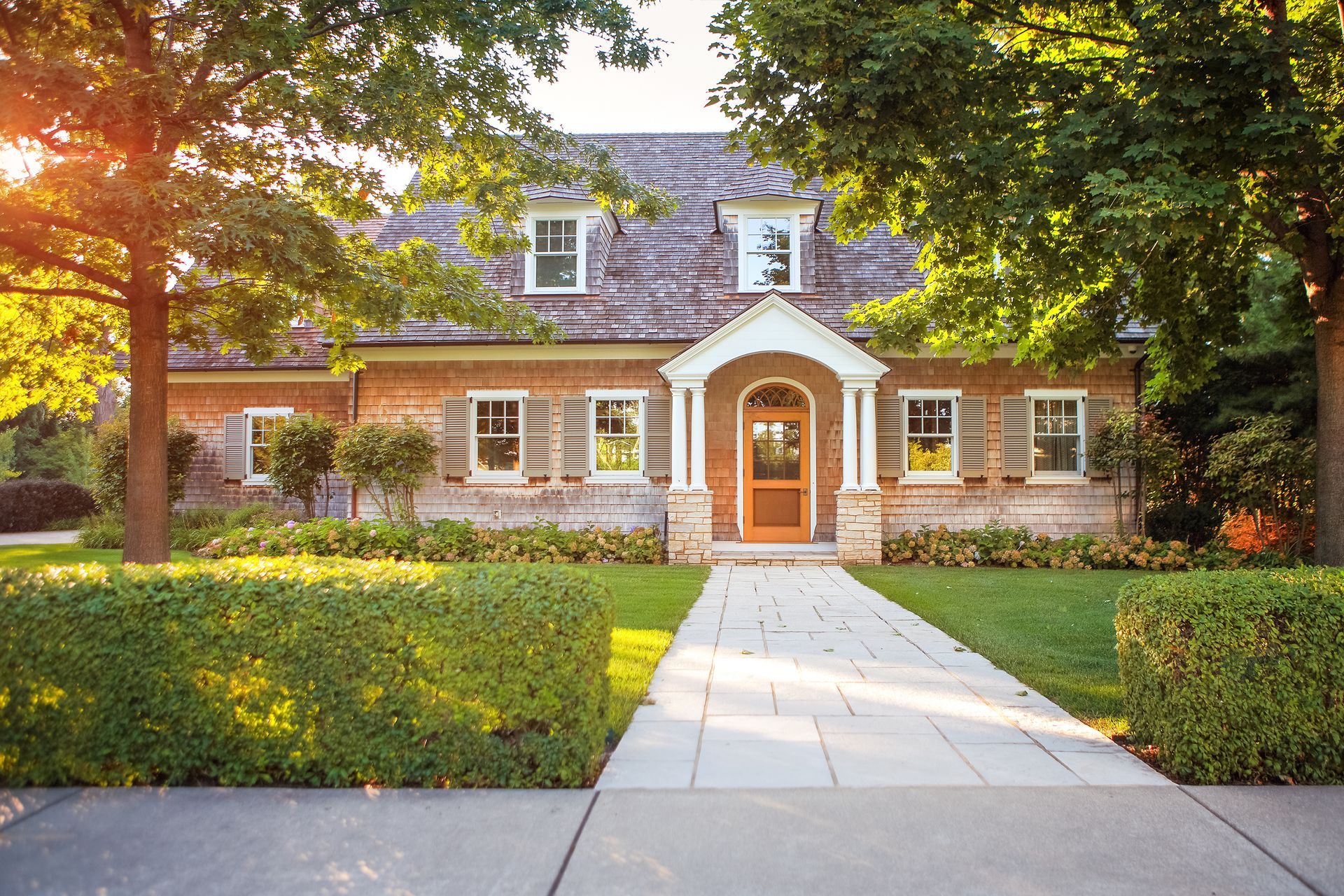 Brick house with white trim and a front walkway, framed by lush green trees and warm sunlight