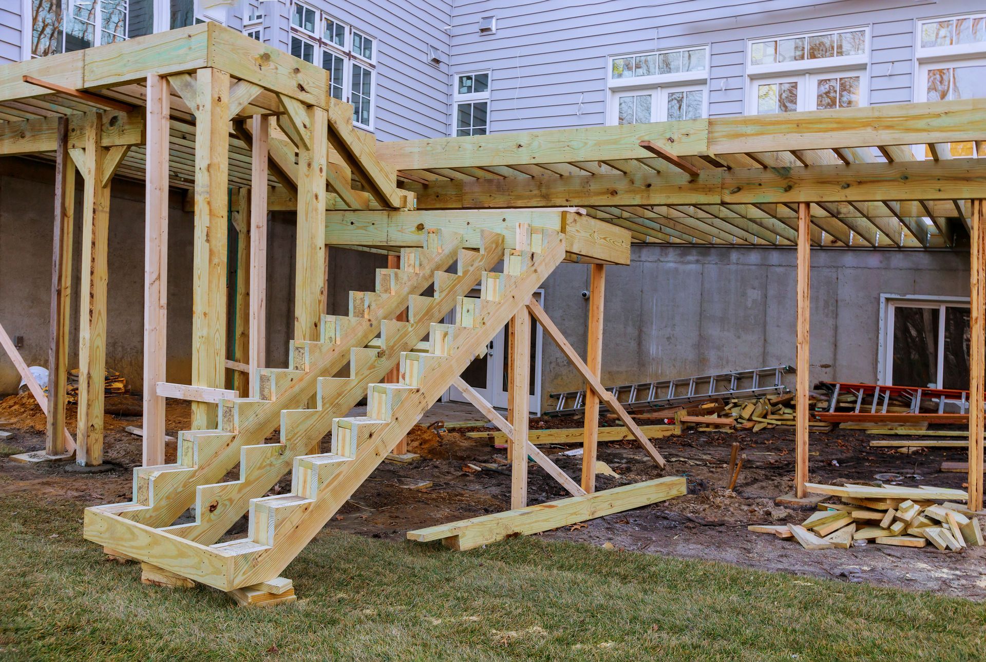 Wooden stairs and deck framing under construction beside a house, with beams, supports, and scattered lumber.