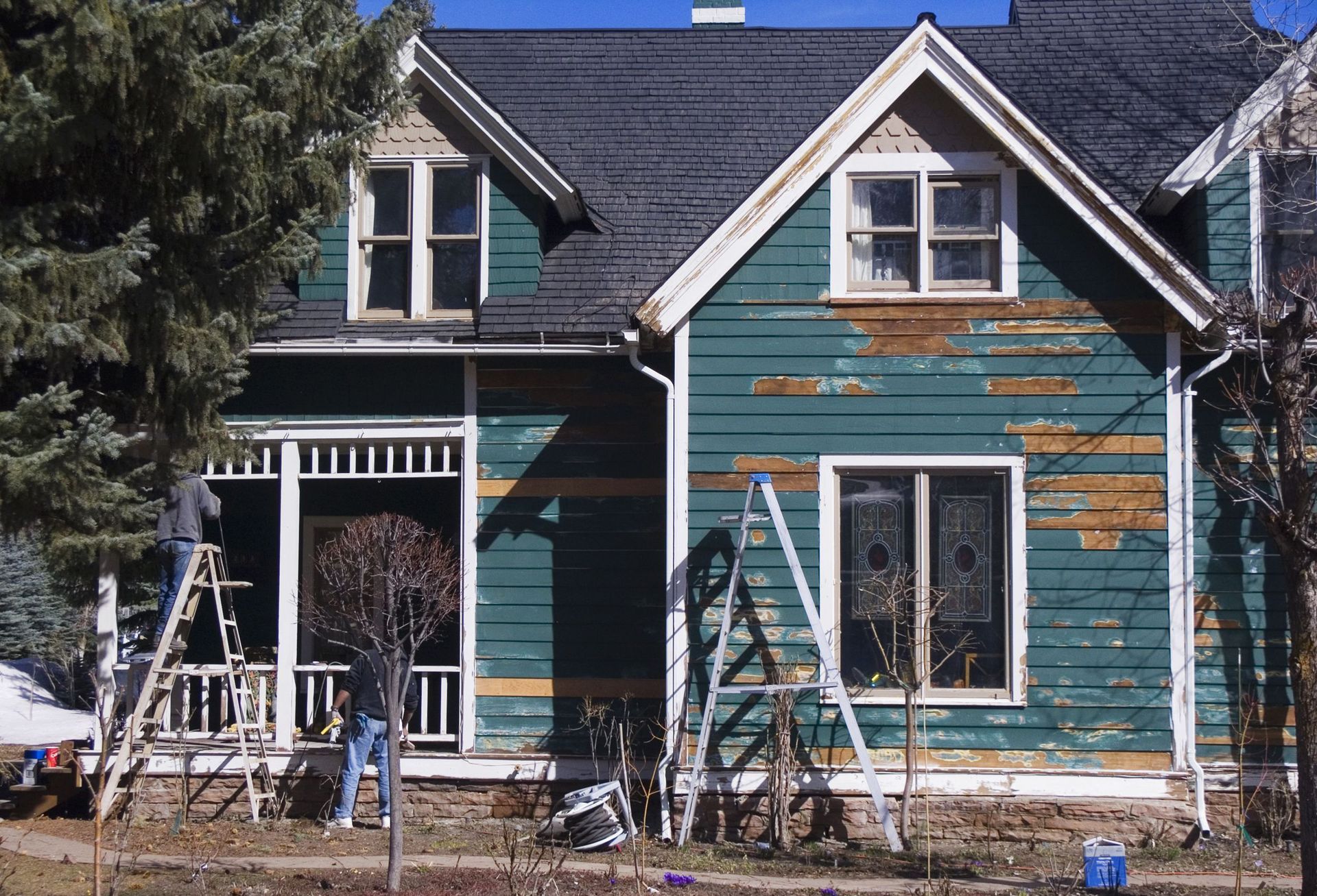 Blue house with peeling paint, front porch, and ladder in a yard under renovation