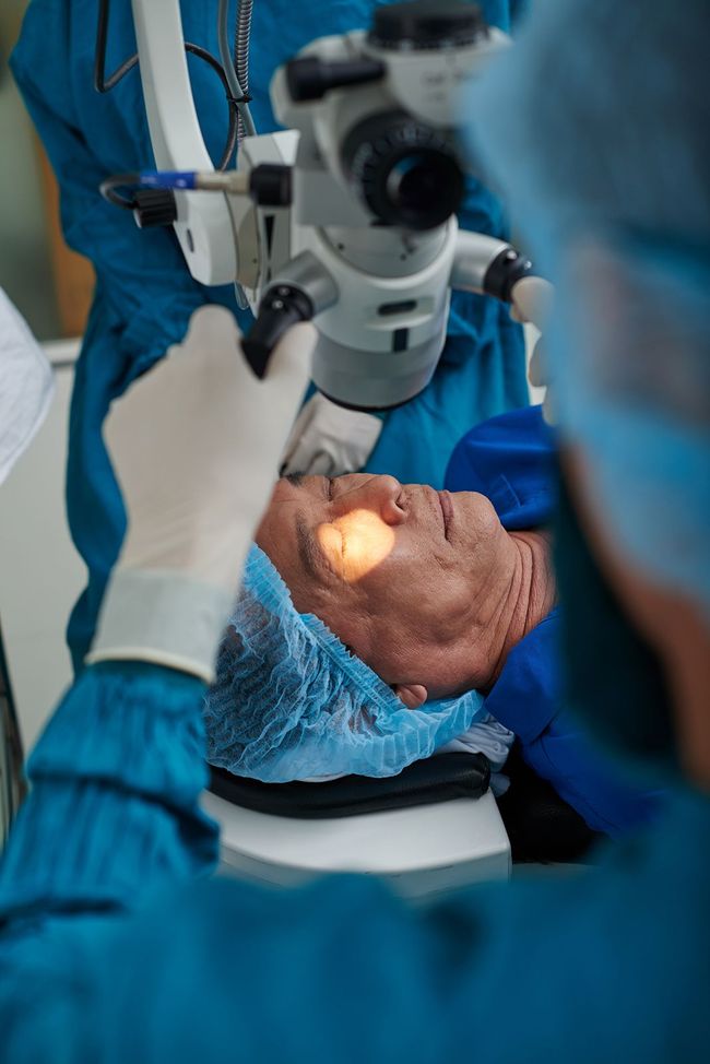 Paciente sometido a cirugía ocular, ojo iluminado. Personal médico con uniforme quirúrgico azul, utilizando un microscopio.
