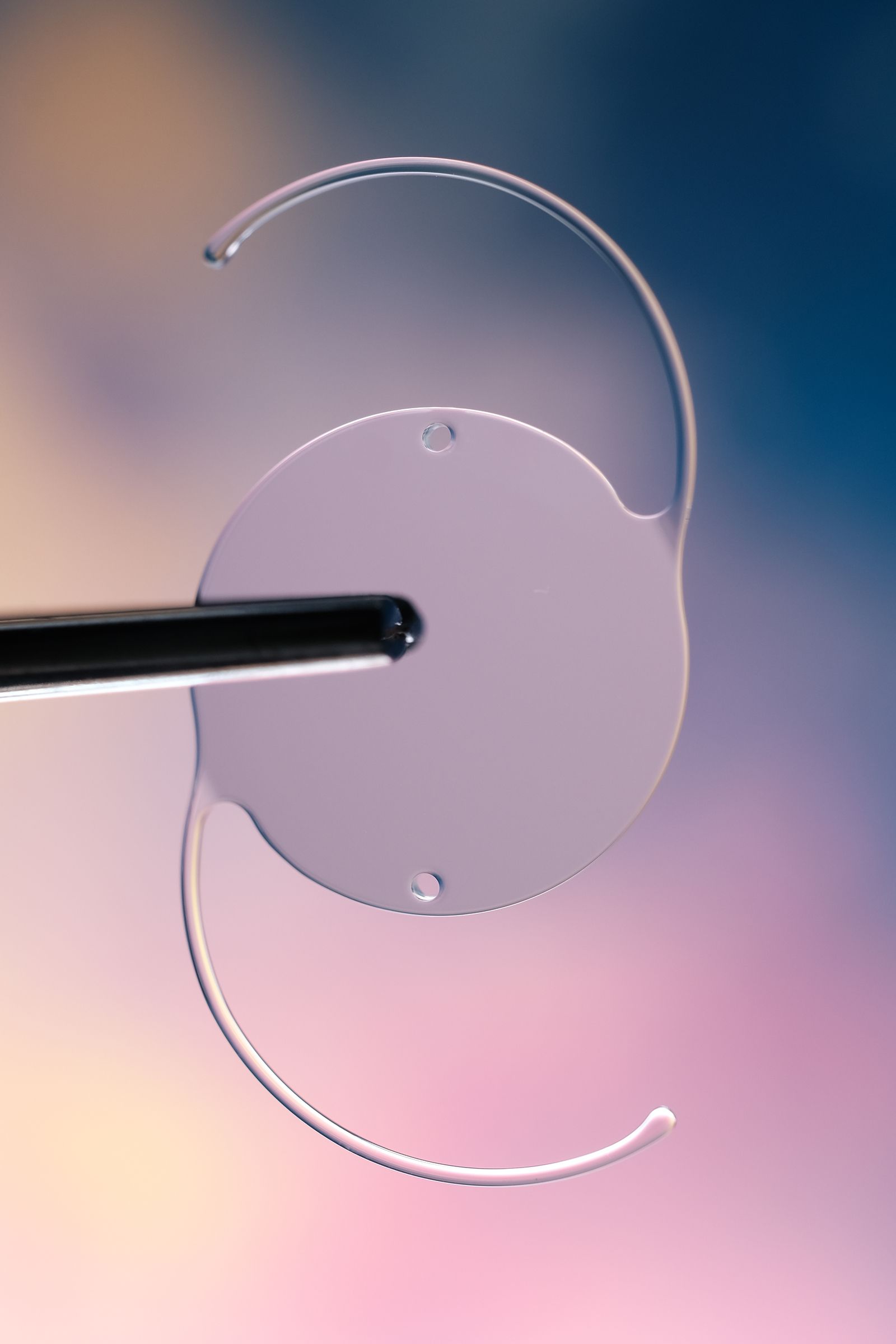 Lente intraocular sostenida con pinza; lente circular transparente con soportes curvos, sobre fondo colorido.