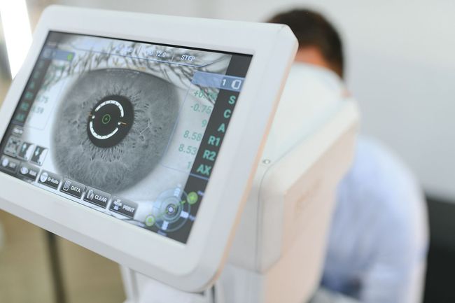 Dispositivo de examen ocular que muestra un primer plano del ojo de un paciente. Una persona aparece desenfocada al fondo.