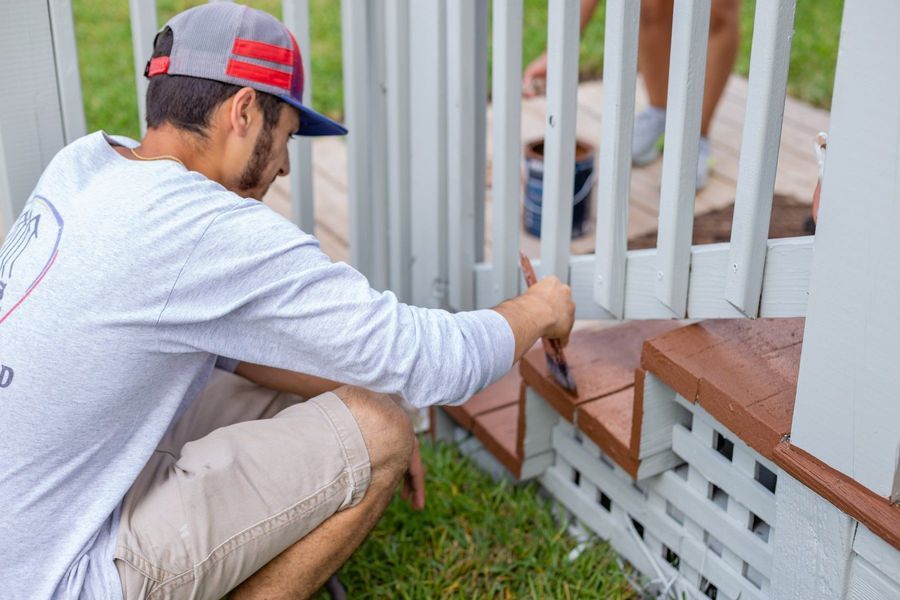 Man in cap painting steps on a deck; brown steps, white railing, green grass.