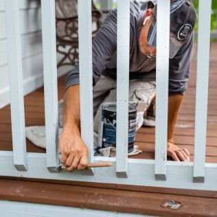 Man paints white railing posts on a porch, holding brush and can, brown deck.