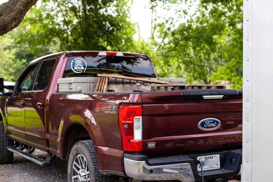 Burgundy Ford Super Duty truck parked outdoors with a toolbox, wood, and a clear window sticker.