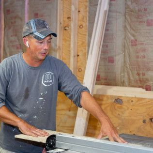 Man in cap using a table saw in a wood-paneled room.