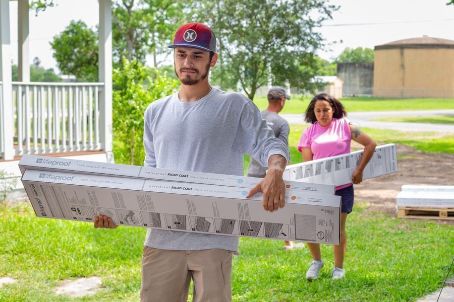 People carrying boxes of flooring in a yard; man in red cap carries several; woman and another man follow.