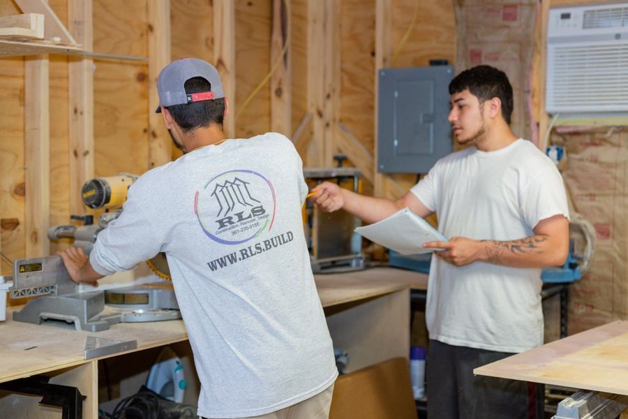 Two men in a workshop. One operates a saw, the other reviews plans and points.