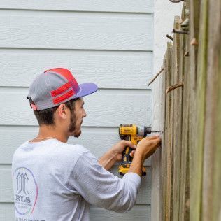 Man in a hat using a yellow drill to attach something to a wooden fence outdoors.